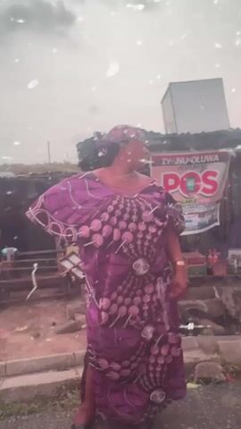 A woman stands in front of a small outdoor business. The woman is wearing a purple patterned dress and headwrap. She has her right hand on her hip and is looking to her right. Behind her, there's a sign that says "IYANU-OLUWA VENTURES POS." The business area looks somewhat cluttered with various items. The weather appears to be overcast, with the presence of rain or some sort of precipitation. The background is a bit blurred, suggesting focus on the woman.