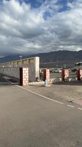The video opens with a view of a paved area with red and white checkered tiles, leading to a dirt path. In the background, there is a fence with red brick pillars and a white wall. Beyond the fence, several parked cars are visible, and further in the distance, mountains stretch across the horizon under a cloudy sky. As the video progresses, the camera pans to the right, revealing a military vehicle displayed on a raised platform. This vehicle is an open-topped jeep with a gun mounted on the front. Another military vehicle, a smaller armored car, is also displayed on a separate platform to the right. The scene appears to be an outdoor exhibition or memorial site. There are no people clearly visible interacting with the vehicles. The environment is dry and dusty, characteristic of a mountainous region.