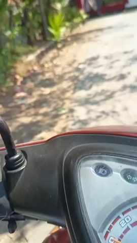 The video shows a first-person view of someone riding a red scooter down a paved road. There are trees and vegetation on the left side of the road, and the ground is covered with scattered debris. The road is partially shaded by tree branches. In the distance, there is an archway with red pillars and a sign that reads "SAMPORES" and "DIRGAHAYU". There are also Indonesian flags hanging from the archway. The overall scene appears to be an outdoor environment, possibly in a village or rural area.