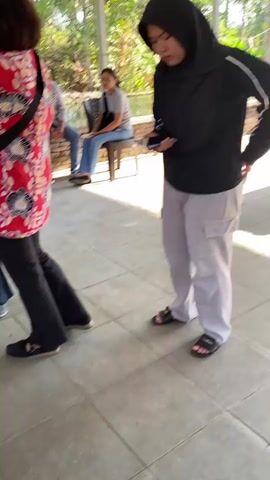 The video shows an outdoor gathering with several people. In the foreground, two women are interacting. One, wearing a black hijab and grey cargo pants, looks down at her phone. The other, in a red and white patterned shirt and black pants, faces away from the camera. Behind them, a woman in a pink hijab speaks to the woman in the red shirt. Further back, several men are seated or standing, some near a table laden with small bowls of food. The environment appears to be a covered patio area with metal roofing, open on the sides to reveal lush greenery and trees. The ground is tiled.
