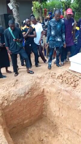 People are gathered around a grave site, which is dug in reddish-brown soil. A white coffin rests beside the open grave, secured by two thin wooden poles placed over the hole. The camera pans to provide a closer view of the grave. Several individuals, dressed in a variety of clothing, are standing at the edge of the grave, observing. Some are wearing casual attire, while others are dressed in more formal clothing. There are trees in the background and a small building to the side.