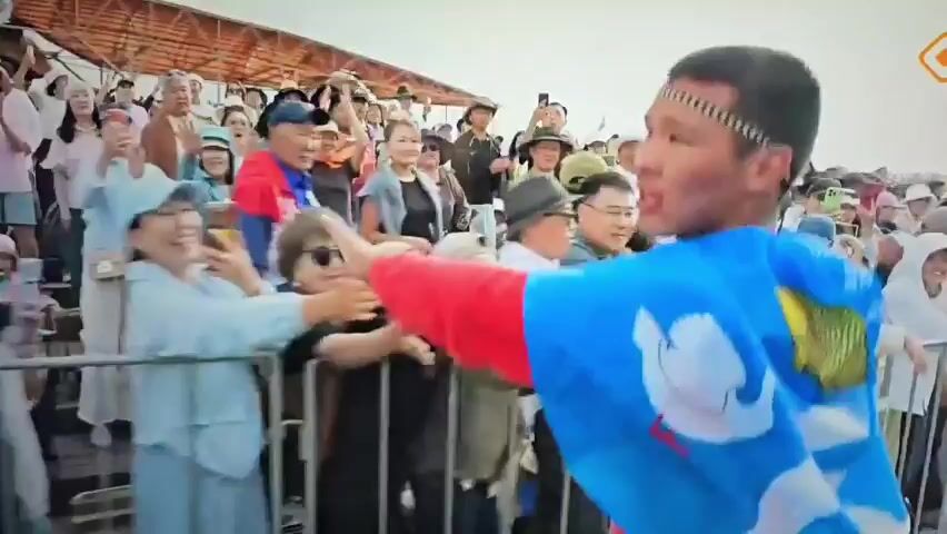 In the first frame, a crowd of people are visible behind a metal fence. Several people are holding up their phones to take pictures. A young person wearing a red shirt and a blue cape is reaching out to the crowd. In the next frames, the scene becomes very dark and blurry, with a faint image of a window with light coming from it. The final frames show the crowd again, with the person in the red shirt and blue cape moving away from the fence and towards the viewer. The person is looking down, and their arms are at their sides. The crowd appears to be cheering and reaching out to the person.