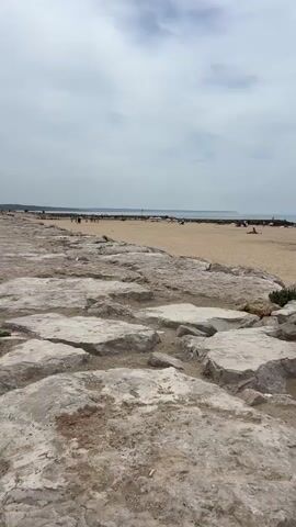 The video opens with a close-up of large, rough rocks in the foreground. The camera pans to the right, revealing a sandy beach with a few scattered rocks and some sparse vegetation. Beyond the rocks, the beach stretches out, dotted with people relaxing under umbrellas, some sunbathing, and others walking. The ocean is visible in the background, with a calm surface and a hazy sky overhead. Several beach umbrellas are set up in rows, with some beach chairs and small tables beneath them. There is a lifeguard stand and a small blue structure on the beach. Further out in the water, a few boats can be seen. The overall atmosphere is one of a relaxed, overcast day at the beach.