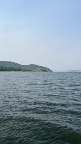 A serene view of a large body of water, likely a lake or sea, under a clear, pale blue sky. Gentle waves ripple across the surface of the water, creating a textured pattern of light and shadow. In the distance, a lush, green shoreline curves into view, dotted with trees that climb the rolling hills. A small, grassy island rises from the water near the shore, adding a focal point to the landscape. The overall impression is one of natural beauty and tranquility.