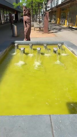 The video shows a rectangular fountain filled with yellowish-green water. Water is being pumped up through several nozzles that create small, clear water streams which then fall back into the pool. The fountain is set on a paved area alongside a street lined with buildings and trees. To the left and right of the fountain stand tall, narrow sculptures made of reddish-brown stone. People and parked motorcycles are visible in the background, moving along the street. The scene is captured under bright daylight.