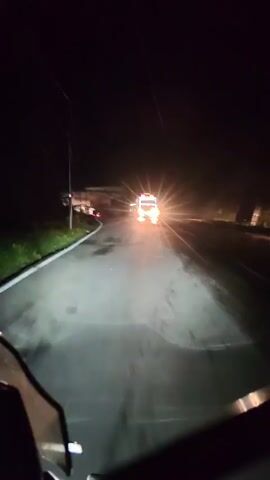 The video begins with a view from a vehicle at night, its headlights illuminating a wet, dark road. In the distance, an ambulance with flashing lights is visible, approaching the camera. As the ambulance gets closer, the camera pans slightly, showing more of the road and the ambulance's illuminated grille. The ambulance passes by, and the camera continues down the road, passing a large white structure on the left side and a bridge or overpass ahead. The video ends with a view of the dark road ahead, with the headlights of the vehicle continuing to light the way.