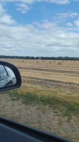 The video shows a view from inside a moving vehicle, looking out the side window. A side mirror reflects the interior of the car and the blue sky with clouds. Outside the car, a harvested field is visible, with rows of hay bales scattered across it. The field extends to a tree line in the distance. The sky is partly cloudy. The vehicle is moving along a dirt or gravel road.