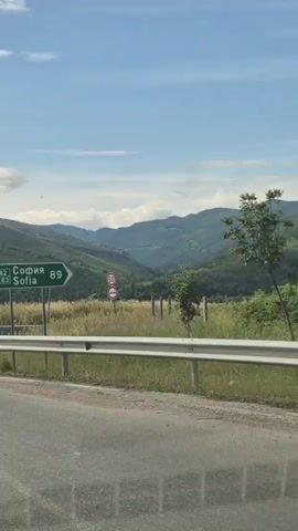 A green road sign indicates Sofia is 89 kilometers away, with the road A2 and E83 also mentioned. The sign is located on the right side of a road that curves to the left. Beyond the road, there is a metal guardrail, followed by a field of tall, dry grass. In the background, rolling green hills are visible under a bright blue sky with scattered clouds. A few traffic signs are also visible. The scene appears to be a scenic rural landscape during the daytime.