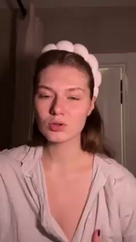 A woman with light brown hair is standing in a dimly lit room, likely a bathroom, wearing a white and light pink striped pajama top and a white plush headband. She is facing the camera and speaking, gesturing with her hands and touching her pajama top. There is a shower curtain rod visible in the background and a closed white door behind her.