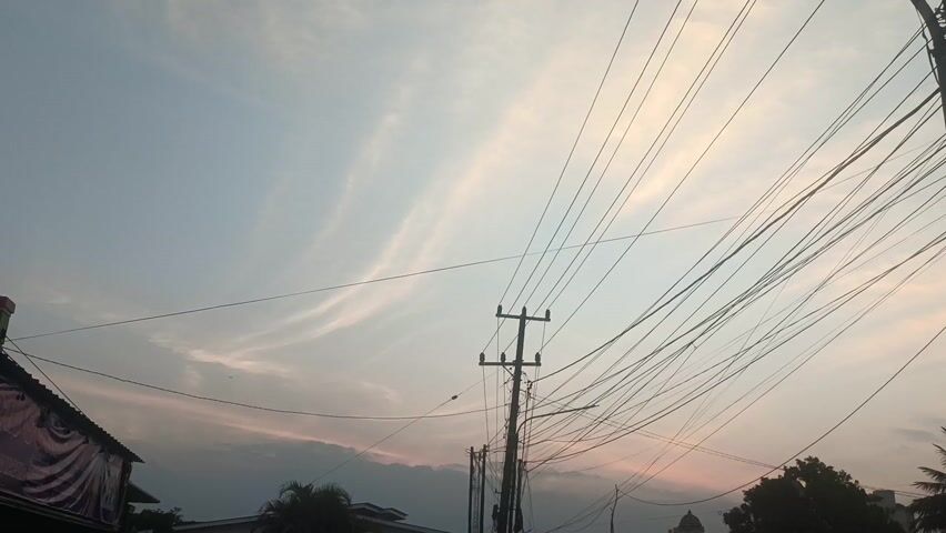 This video captures a street scene in Sungailiat, Indonesia, during what appears to be sunset. The sky is a soft gradient of pale blue and pink, with wispy clouds streaking across the frame, indicating a partly cloudy evening.

The foreground is dominated by a dense network of overhead power lines, strung between numerous utility poles. These wires crisscross the sky, creating a complex, almost abstract pattern against the twilight.

On the street level, the scene is populated with various vehicles and structures. Several cars, including a white minivan with its doors open, and a yellow hatchback, are visible, suggesting moderate traffic. A few motorcycles can also be seen, with riders evident on some.

Buildings line the street, with one on the left featuring a dark roof and a banner with indistinct text. Further down the road, the silhouette of a building with a dome is discernible against the sky, hinting at a religious or civic structure.

The general atmosphere is that of a bustling yet calm evening in a town. The soft light of sunset casts a gentle glow over the scene, creating a somewhat tranquil mood despite the electrical infrastructure. The time of day is clearly evening, as indicated by the sky's coloration and the ambient light. There are no strong weather elements apparent, suggesting fair weather.

Visible text is minimal and mostly illegible due to distance and resolution, but a sign on a shop appears to have Indonesian writing.