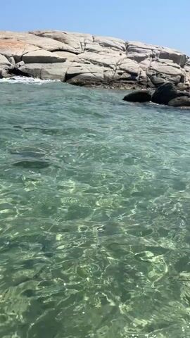 The video shows a clear, shallow body of water with sunlight creating rippling patterns on the surface. In the background, a rocky shoreline with some vegetation is visible under a clear blue sky. A few people are standing on the rocks in the distance. The camera appears to be moving slightly, suggesting it is handheld or on a boat, and it focuses on the water and the rocky landscape. The overall scene depicts a tranquil coastal environment on a sunny day.