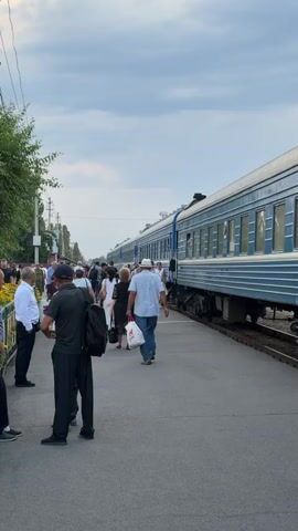 The video shows a train at a station platform with many people on it. The train is long and blue with white stripes. The people are dressed in casual clothing and many are carrying bags, suggesting they are passengers. Some people are walking along the platform, while others appear to be waiting to board or disembark. The scene is set outdoors, with trees and utility poles visible in the background. The sky is cloudy, indicating it could be daytime, possibly late afternoon or early evening. The overall atmosphere appears to be one of a busy train station, with people moving about. There is no visible text in the video.
