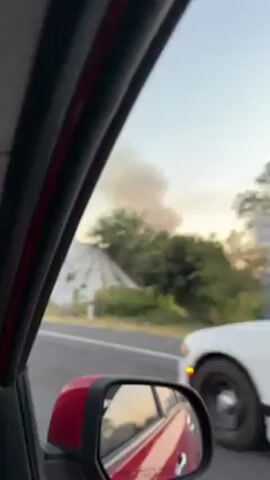 The video shows a car driving on a highway. Out of the car window, a plume of dark smoke rises into the air in the distance, against a sunset sky. The camera passes under a concrete overpass. Trees and vegetation are visible along the side of the road. The driver’s side mirror of the car is visible in the lower left corner.