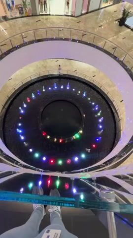 A high-angle, vertical shot shows a circular fountain with colorful lights in the center of a multi-level shopping mall. The fountain has several jets of water spraying upwards, illuminated by blue, red, and green lights. A person stands on the edge of the fountain, looking at it. The mall has several levels with shops visible in the background. The viewer's feet and legs are visible at the bottom of the frame, suggesting they are standing on a balcony overlooking the fountain.
