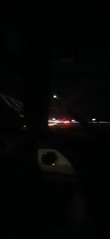 The video captures a nighttime view from inside a vehicle moving along a road. The foreground is dark, with a portion of the dashboard or car interior visible in the lower part of the frame. The windshield shows streaks, possibly from rain or dirt. Through the windshield, streaks of light from oncoming traffic and streetlights are visible, creating a blurred, streaky effect due to the vehicle's motion and low light conditions. The overall impression is a fast-moving perspective on a highway or road at night.
