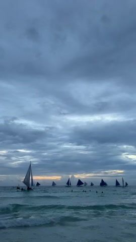 The video shows a wide shot of a beach at dusk. The sky is filled with dark, dramatic clouds, with a sliver of orange visible at the horizon. The ocean is a choppy, deep blue, with white waves breaking near the shore. In the distance, several paraw sailboats, with their distinctive triangular sails, are visible on the water. Some people are in the water near the shore. The scene is serene and slightly ominous due to the dark clouds.