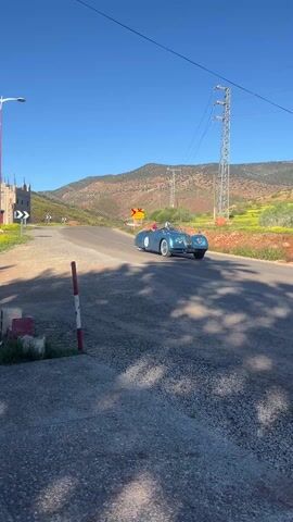 A blue vintage race car drives on a paved road towards the camera, passing through an intersection marked by road signs. The environment includes hills, yellow fields, utility poles, and scattered buildings under a clear blue sky. A person in a helmet is visible in the car. The foreground shows a rough, gravelly surface with a post and concrete blocks.