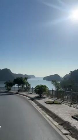 The video shows a scenic view of a bay with several small islands in the distance. The water is calm and reflects the bright sunlight. In the foreground, there's a road with a pedestrian walkway lined with a metal fence. Small trees are planted along the walkway. A large white boat is visible on the water between two of the islands. The sky is clear and blue.