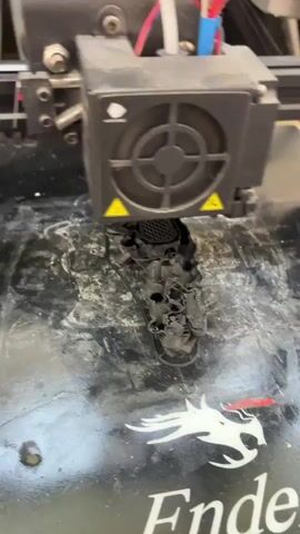 This media file captures a close-up view of a 3D printer in operation. The primary object is the print head of the 3D printer, which is actively depositing black filament onto a build plate. The filament appears to be melting and solidifying as it's laid down, forming a complex, possibly intricate, object. There is some white material on the build plate, which might be residue from previous prints or a cleaning agent.

The scene is illuminated by artificial light, suggesting it could be any time of day. The background is out of focus but appears to be an indoor setting, possibly a workshop or a hobbyist's space. The build plate itself has a distinctive design, featuring a white dragon logo and the partial text "Ender" prominently displayed. This suggests the printer is likely an Ender model, a popular brand among 3D printing enthusiasts.

The focus is entirely on the printing process. The 3D printer is the sole subject, and the activity is the creation of a physical object layer by layer. There are no people visible, nor any indication of the weather or specific time of day beyond the artificial lighting. The emotion conveyed is one of focused activity and technological creation. The location context of Toruń, Poland, is an external piece of information not directly visible in the media itself but noted for this description.