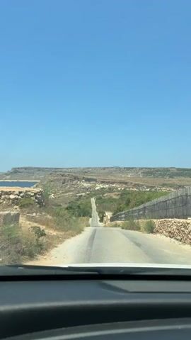 A drone shot shows a straight, paved road leading towards a rugged, rocky landscape under a clear blue sky. The road is bordered by dry brush and a wooden fence. In the distance, the landscape appears arid with sparse vegetation and a body of water visible on the left. A person can be seen walking down the road in the distance. The view is from a car, as indicated by the dashboard at the bottom of the frame.
