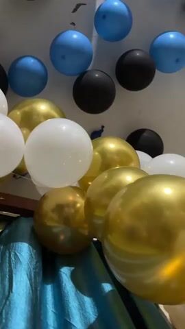 A close-up, low-angle shot shows a cluster of inflated balloons. The balloons are of various colors: white, gold, black, and blue. Some are higher up, attached to what appears to be a ceiling or upper wall, while others are closer to the foreground, resting on a draped fabric that is teal and green. The lighting is somewhat dim, and the balloons are reflecting the light. The focus shifts slightly throughout the short clip, creating a dynamic view of the balloon arrangement. No people are visible.