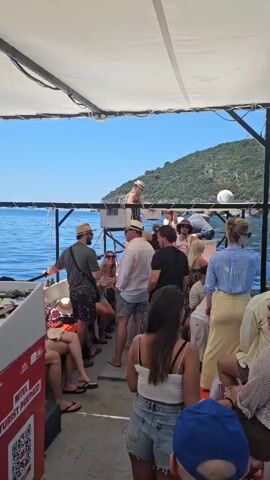 A group of people are gathered on the deck of a boat, likely on a tour or excursion. The boat is moving through the water, indicated by the slight blur of the background and the presence of the sea and a green, hilly coastline. Many of the individuals are wearing summer attire, including hats, shorts, and light-colored shirts. Some are standing and talking, while others appear to be seated or leaning. One person is standing on a raised platform near the center of the boat, facing away from the camera. The overall atmosphere seems relaxed and social, typical of a daytime boat trip.