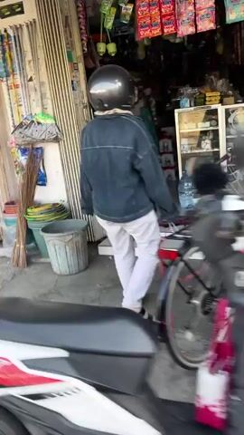 The video shows a person wearing a black helmet and a denim jacket, white pants walking into a store. A bicycle is parked next to the store. In the foreground, part of a motorcycle is visible. The store appears to be a small retail establishment with shelves stocked with various goods. There are also bins and brooms outside the store.