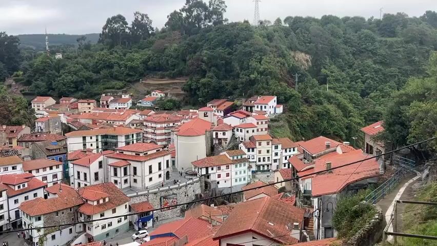 The video shows a panoramic view of a densely packed village nestled in a valley. The buildings are predominantly white with red tile roofs and are clustered together on the slopes of a tree-covered hill. A cylindrical structure, possibly a tower or church, stands prominently in the center of the village. There are a few people visible walking on pathways and in small plazas within the village. The sky is overcast, suggesting a cloudy day. The overall impression is of a traditional, somewhat historic European coastal village.