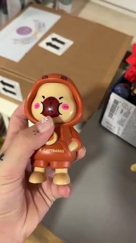 A person's hand holds a small, orange, cartoon-like toy figure with a hood. The toy has painted features on its face, including eyes, cheeks, and a mouth. The toy's face animates with blinking eyes and mouth movements. In the background, there is a cardboard box with stickers and other items on a surface. The hand holding the toy has a tattoo on the thumb and a painted flower design on one fingernail.