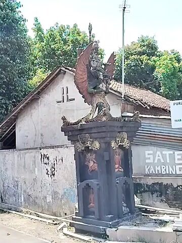 The video shows a black, ornate statue of Garuda, a mythical bird-like creature from Hinduism, on a pedestal. The statue is located near a building with a tiled roof and trees in the background. The statue depicts Garuda with large wings, carrying a figure on its back. There are some decorative elements on the pedestal beneath the statue. The scene is well-lit, suggesting it is daytime. The camera appears to be at eye level, possibly handheld.  A tall, metal antenna is visible in the background as well.