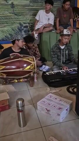 The video shows a group of young men in what appears to be a casual gathering, possibly a rehearsal or a performance, indoors. The environment features a tiled floor and a wall decorated with a mural of what looks like terraced rice fields. In the foreground, there is a large, ornate wooden percussion instrument, possibly a Gamelan instrument, on a stand. A young man wearing a black t-shirt and a patterned cap is seated to the left, holding a cigarette and looking at the instrument. Next to him, another young man in a patterned shirt is looking at his phone. To the right, a young man in a camouflage-patterned hoodie is seated in front of an electronic keyboard, labeled "MEDELI", and appears to be playing it. In the background, two other young men are seated on a raised platform or bench. One, wearing a white t-shirt, is looking towards the side, while the other, wearing a brown t-shirt, is looking down. There are several objects scattered on the floor, including a cardboard box containing cups, a silver thermos, and another white box with red markings. The lighting is adequate, suggesting an indoor setting with artificial light. The overall atmosphere seems relaxed and informal.
