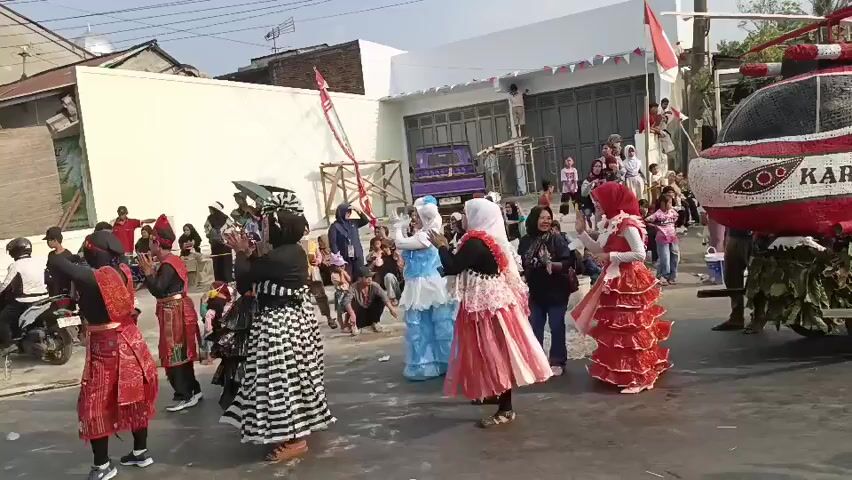 A group of people are gathered in the street, some wearing colorful traditional clothing and others in casual attire. Several individuals appear to be performing a dance, with synchronized movements and clapping. A decorated float shaped like a helicopter is visible on the right side of the frame, adorned with red and white elements. The event seems to be a celebration or parade, indicated by the festive atmosphere and the presence of onlookers. The background shows a street lined with buildings, suggesting an urban or suburban setting. The Indonesian flag is displayed prominently on a pole behind the helicopter float.