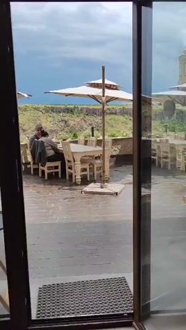 The video shows an outdoor patio area with tables and chairs, likely a restaurant setting. Two people are seated at a table on the left side of the frame, facing away from the camera. They appear to be conversing. A large umbrella stands over the table where the two individuals are seated. In the background, there is a view of a rocky landscape with some vegetation. The ground of the patio is paved with stone tiles and appears wet, suggesting it may have recently rained. The sky is cloudy. The camera appears to be positioned inside a doorway, looking out at the patio.