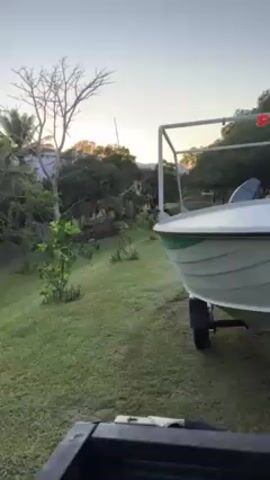 The camera is mounted on the back of a vehicle, likely a truck. The vehicle is parked on a grassy lawn. A boat on a trailer is visible to the right. The boat is white with a green stripe. It has a metal frame structure on top. In the background there are trees and mountains visible under a light blue sky. The time of day appears to be early morning or late afternoon.