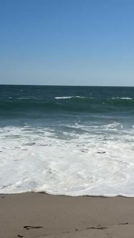 The video shows a tranquil ocean scene with gentle waves lapping against a sandy beach. The water is a deep blue-green, and the sky is a clear, bright blue. The waves create a foamy white surf as they break on the shore. The sand appears light brown and undisturbed, except for a few faint footprints. The overall impression is one of peaceful serenity and natural beauty.