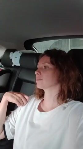 The video shows the interior of a car from the perspective of someone in the front passenger seat, looking back. A woman in a white t-shirt is in the foreground, partially blocking the view. In the back seat, a young girl is sitting in a car seat, holding a tablet and a phone. A bottle of water and a portable charger are also visible on a tray in front of her. The car appears to be in motion, with trees and a road visible through the windows. A stuffed animal is perched on the headrest of the driver's seat.