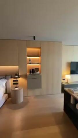 The video shows a panning view of a modern hotel room. The floors are wooden, and the walls are paneled with light-colored wood. There is a built-in shelving unit on the left wall with an illuminated display niche. In front of it is a small round table with items on it. A low, dark brown entertainment center sits along the wall. Above it is a flat-screen television. In the center of the room, there is a large sectional sofa and two round tables. A white chair and a floor lamp sit in front of a large window. Beige and white curtains cover the window. The room is well-lit. The overall style of the room is modern and minimalist. There is a bottle of water and two glasses on a small table in the foreground.