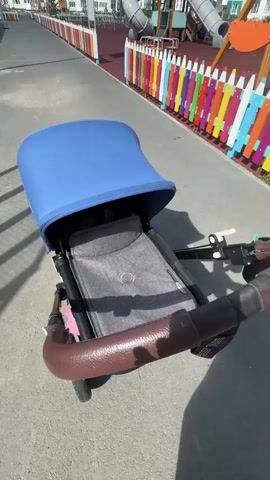 The video shows a stroller with a blue canopy and a gray seat. The stroller has a brown handle. In the background, there is a playground with colorful fences and slides. Several tall buildings are visible behind the playground. A young woman wearing sunglasses and a light blue t-shirt is pushing the stroller. She is looking to the side and smiling. The scene is outdoors on a sunny day.