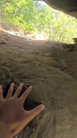 The video shows a first-person perspective of someone walking down a steep, muddy incline. The path is rough and uneven, with loose dirt and rocks. Green foliage and trees are visible at the top of the incline, suggesting an outdoor, natural environment. The lighting indicates it is daytime, possibly overcast or dappled sunlight filtering through trees. The individual is moving downwards, with the camera angled towards the ground and the slope ahead. There are no other people or objects clearly identifiable in the scene apart from the natural terrain.