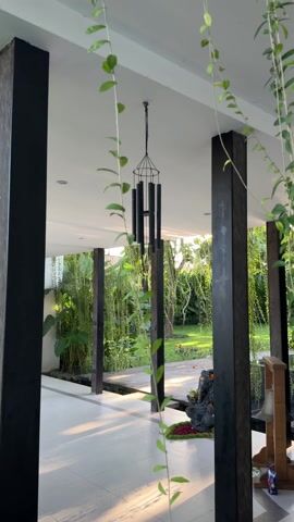The video captures a view of a covered porch or entrance area that opens onto a lush green garden. The foreground is framed by two vertical black pillars and hanging vines with small green leaves. In the center hangs a black tubular wind chime suspended from the ceiling. Beyond the pillars, there is a stone or tile walkway that leads to a wooden boardwalk extending into the garden. A statue of a seated figure, possibly a deity or a Buddha, sits on a small patch of greenery near the walkway. The garden features various trees and plants, some with trailing vines. The lighting suggests it is either early morning or late afternoon, with soft light casting shadows on the floor. There is also a small shelf or stand with a bell and a few items on it. The scene is tranquil and evokes a sense of peace and nature.