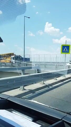 This is a video of a large yellow concrete pump truck driving down a road. The truck is white on the front with a yellow boom extending from the back. The truck is passing a pedestrian crossing sign and a metal guardrail. There are white clouds in the blue sky.