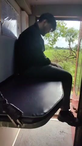 A man in a black shirt and gray pants sits on a dark-colored seat, his head bowed and his eyes closed, appearing to be asleep. He is facing the open doorway of what looks like a train car, with lush green trees visible outside. The seat is an angled, padded bench, with metal brackets at the bottom. The interior of the train car is dimly lit, with a poster or map visible on the wall behind the man. The camera is positioned to capture the man from the side, with the focus on his resting posture and the backdrop of the passing scenery.