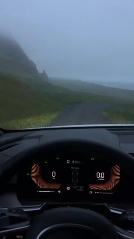 The video presents a driver's perspective from inside a car traveling on a gravel road in a misty, mountainous landscape. The car's dashboard is visible, showing a digital display indicating "Electric mode," a speedometer registering between 6 and 15 km/h, and a range of 420 km. The weather appears overcast and drizzly, with water droplets on the windshield. The surrounding terrain is mostly green with grassy slopes and rocky formations, disappearing into a thick fog in the distance.
