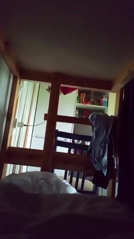 The video appears to be filmed from a low angle, possibly from a bed, looking up at a bunk bed frame. The lighting is dim, with a spotlight effect on some parts of the bunk bed. The bunk bed is made of wood and has a ladder. Some clothing is hanging on the bunk bed. In the background, there is a shelf with various items and a white appliance, possibly a refrigerator or a cabinet. The scene appears to be indoors, in a bedroom. The camera movement is slightly shaky and disorienting.