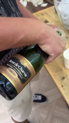 A person is pouring champagne from a bottle into a glass. The bottle is green with a gold label that reads "Brut de Charvis Brut de Traditionnelle." The person is wearing a black and patterned top and white shorts or skirt. They are also wearing black and white sneakers. The champagne is being poured onto a wooden table, which is also holding a bowl of ice and a small white cup. The background is blurred, but appears to be an indoor or outdoor setting with floral decorations. The lighting is bright and natural.