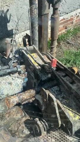 The video shows a close-up view of the working mechanism of a drilling machine, heavily soiled with mud and grease. The machinery consists of hydraulic components, tubes, and metal framing in various states of rust and paint. A red plastic stopper is visible on one of the metal pieces. The machine is actively drilling, as evidenced by the movement of the components and the emission of steam. A concrete wall with patches of exposed brick is visible in the background, along with some weeds and grass. The shadow of a person can be seen cast on the wall, presumably the individual operating the machinery.