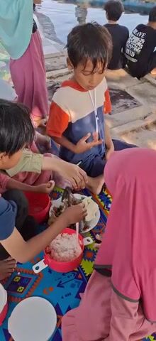 A group of children are sitting on a colorful mat on the ground, eating. The mat is spread out near a body of water, possibly a lake or river. One boy in a blue and orange shirt is looking at the food. Another boy in a blue shirt is eating with his hands. A girl wearing a pink hijab and another girl wearing a light blue hijab are also present and eating. There are several containers of food, including rice and what appears to be fish. The children are casually dressed and seem to be enjoying their meal outdoors.