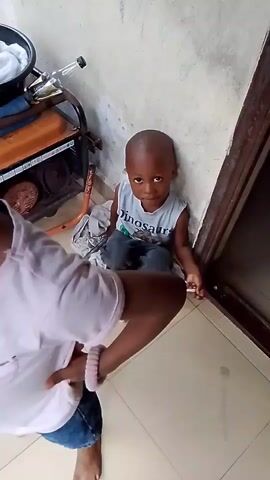 The video shows two young children. One child, a boy, is sitting on the floor, leaning against a wall, wearing a white tank top with "Dinosaur" written on it and blue pants. He looks at the camera with a neutral expression. The second child, a girl, is in the foreground, facing away from the camera, wearing a light pink t-shirt and blue shorts. She has her back to the camera and her hair is styled in braids. The environment appears to be indoors, with tiled flooring and a wall with some graffiti or marks on it. To the left of the boy, there is a generator and some other objects. The girl is blurred in the foreground as she moves.