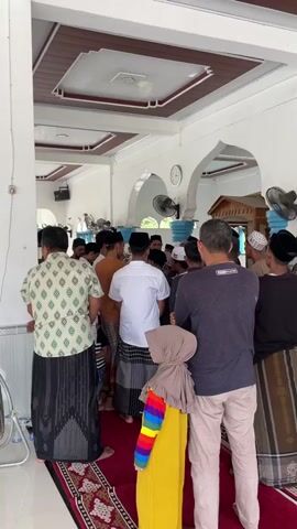 The video shows a group of people gathered indoors, possibly in a mosque, as indicated by the traditional Islamic clothing worn by many of the men. The architecture features white columns and arched doorways, with a wooden ceiling. A red and black patterned rug covers the floor. In the foreground, a person wearing a dark t-shirt and light-colored pants faces away from the camera. To their left, a child in a yellow dress with colorful stripes and a headscarf is also visible. A young child in a striped shirt and jeans is walking on the rug. The majority of the individuals are men, many wearing sarongs and peci (caps). Some are engaged in conversation, while others appear to be observing or waiting. Fans are mounted on the ceiling, suggesting a warm climate. The lighting is bright, indicating daylight. The overall atmosphere seems to be one of a social gathering or a religious event.