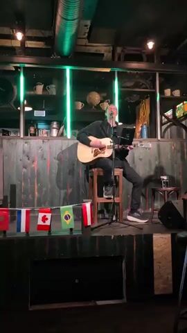 A man sits on a stool on a stage playing an acoustic guitar and singing. He is wearing a black hoodie and black pants. The stage is decorated with flags from different countries. There are green lights above the stage. Behind the man is a bar area with shelves of bottles and mugs. There is a microphone in front of him and a laptop on a stand to his right. A speaker is visible to the right of the stage. The background is made of dark wooden planks.